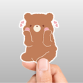 Cartoon bear sticker held between fingers against a gray background