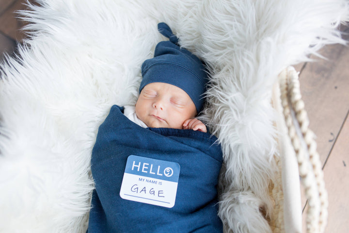 Newborn baby wrapped in blue swaddle with name tag, lying on fluffy white surface