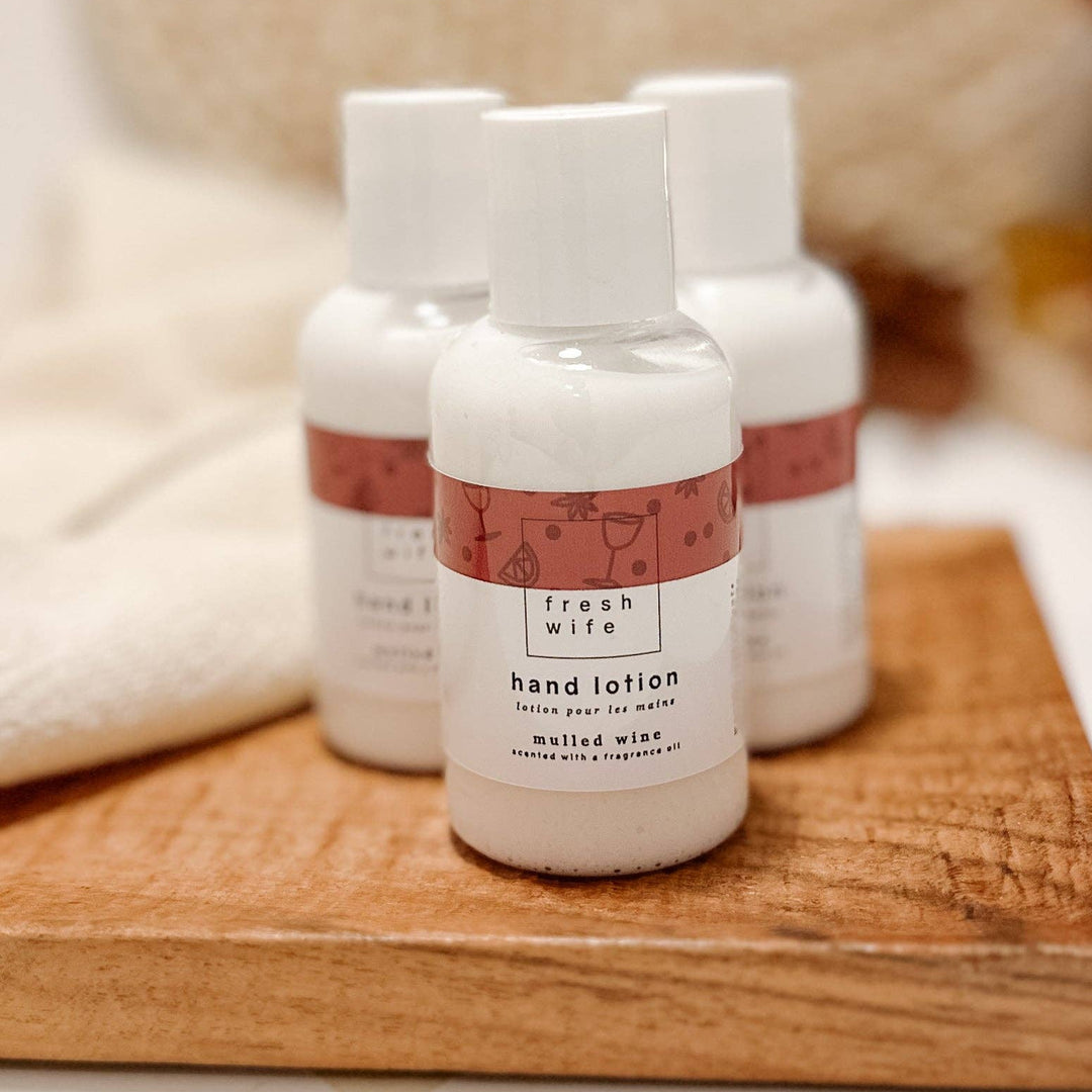 Four small white hand lotion bottles with white caps and pink labels are arranged on a wooden board. The front bottle is in sharp focus, while the others are softly blurred in the background.