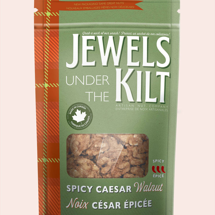 A stand-up pouch of Jewels Under the Kilt Spicy Caesar Walnut, featuring a sage green background with a red-orange tartan pattern on the left side and a clear window showing the walnut pieces inside.
