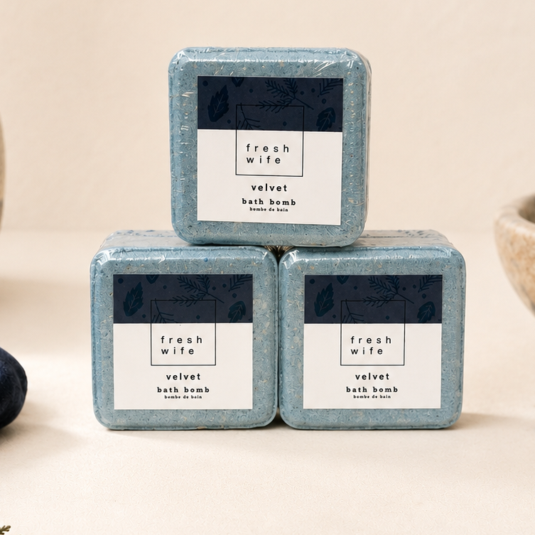 Three square bars of Fresh Mink Velvet Bath Bomb stacked in a pyramid arrangement on a beige surface, styled with dried flowers, a blue velvet cloth, and a bowl of blueberries.