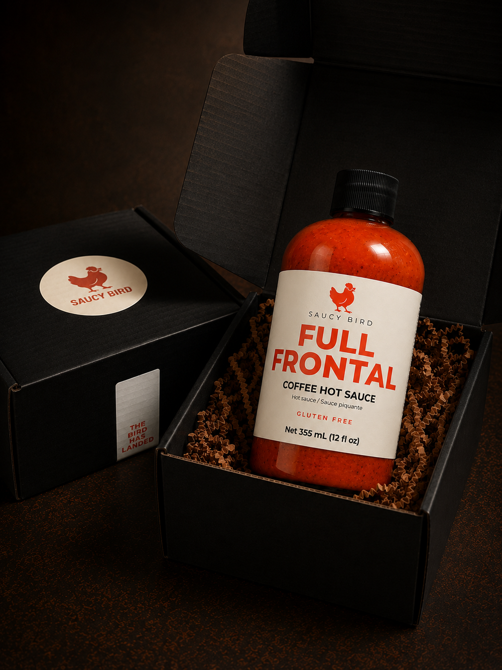 A bottle of Salty Bird Full Frontal Coffee Hot Sauce displayed in an open black gift box with shredded brown paper filler, alongside a branded black box lid.