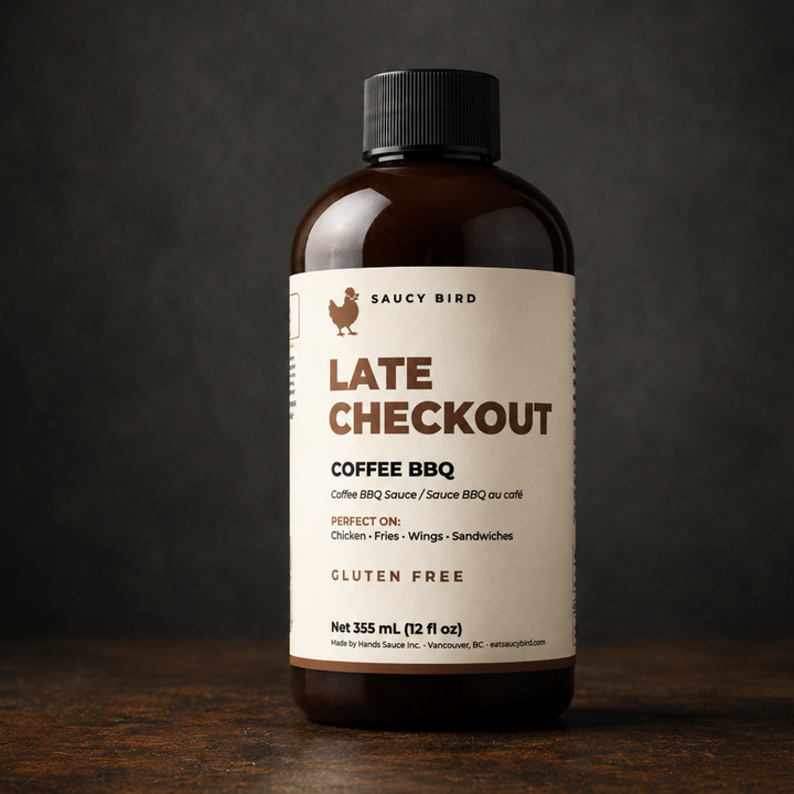 A 355 mL amber glass bottle of Saucy Bird Late Checkout Coffee BBQ sauce, featuring a cream-colored label with brown text, set on a dark wooden surface against a dark gray background.