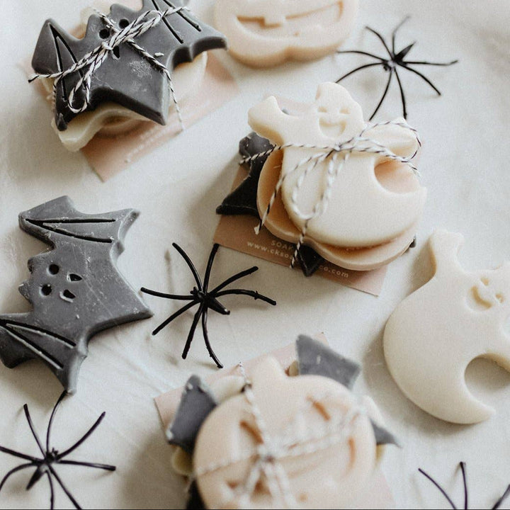 Halloween-themed soap bars shaped like bats, ghosts, and pumpkins, tied with twine and displayed on a light fabric background with black spider decorations.