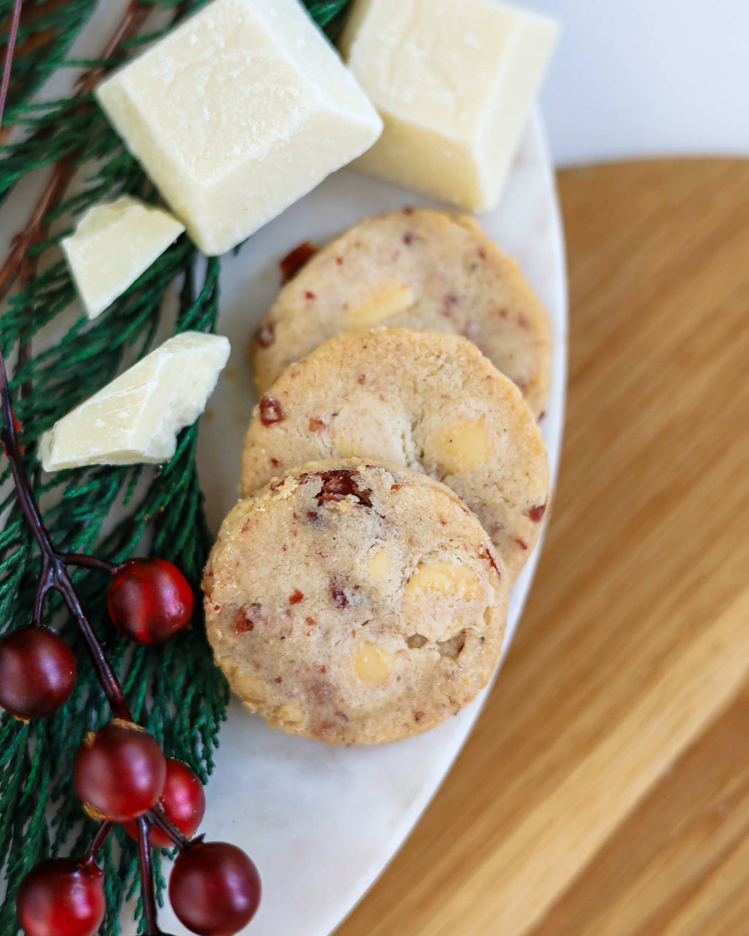 Three round shortbread cookies with nuts and dried fruit, served on a white plate with white chocolate chunks, evergreen branches, and red berries on a wooden surface.
