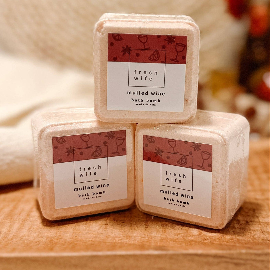 Three square bath soaks in light beige packaging with white and dusty red labels, stacked on a wooden board with a softly blurred spa-like background.