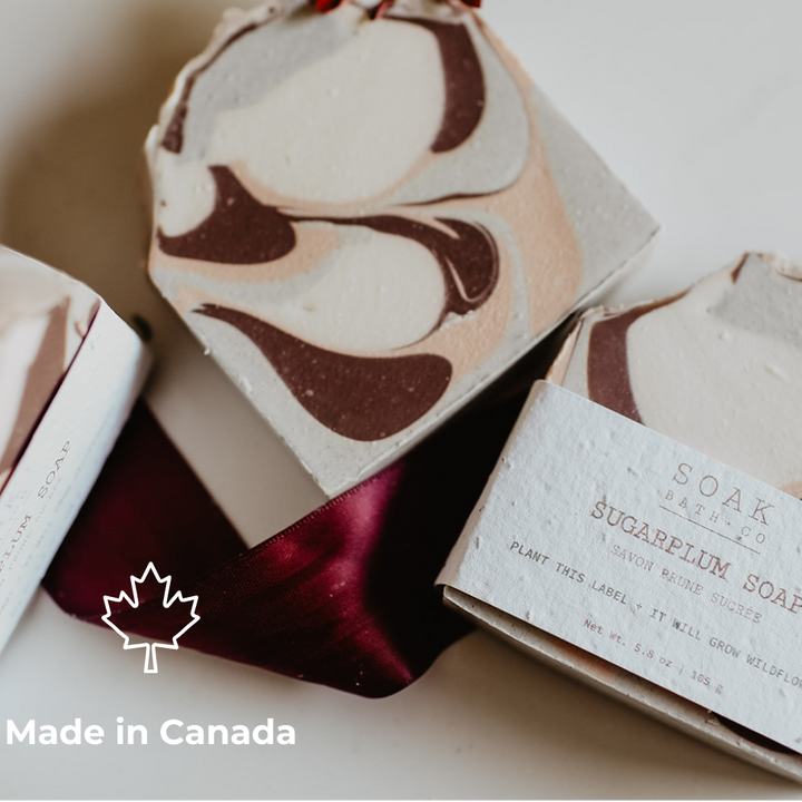 A close-up of handmade soap bars with a marbled cream, beige, and dark brown swirl pattern, wrapped in textured paper labels. One bar is topped with a small red flower, and a white ribbon with a dark red sheen is draped underneath.