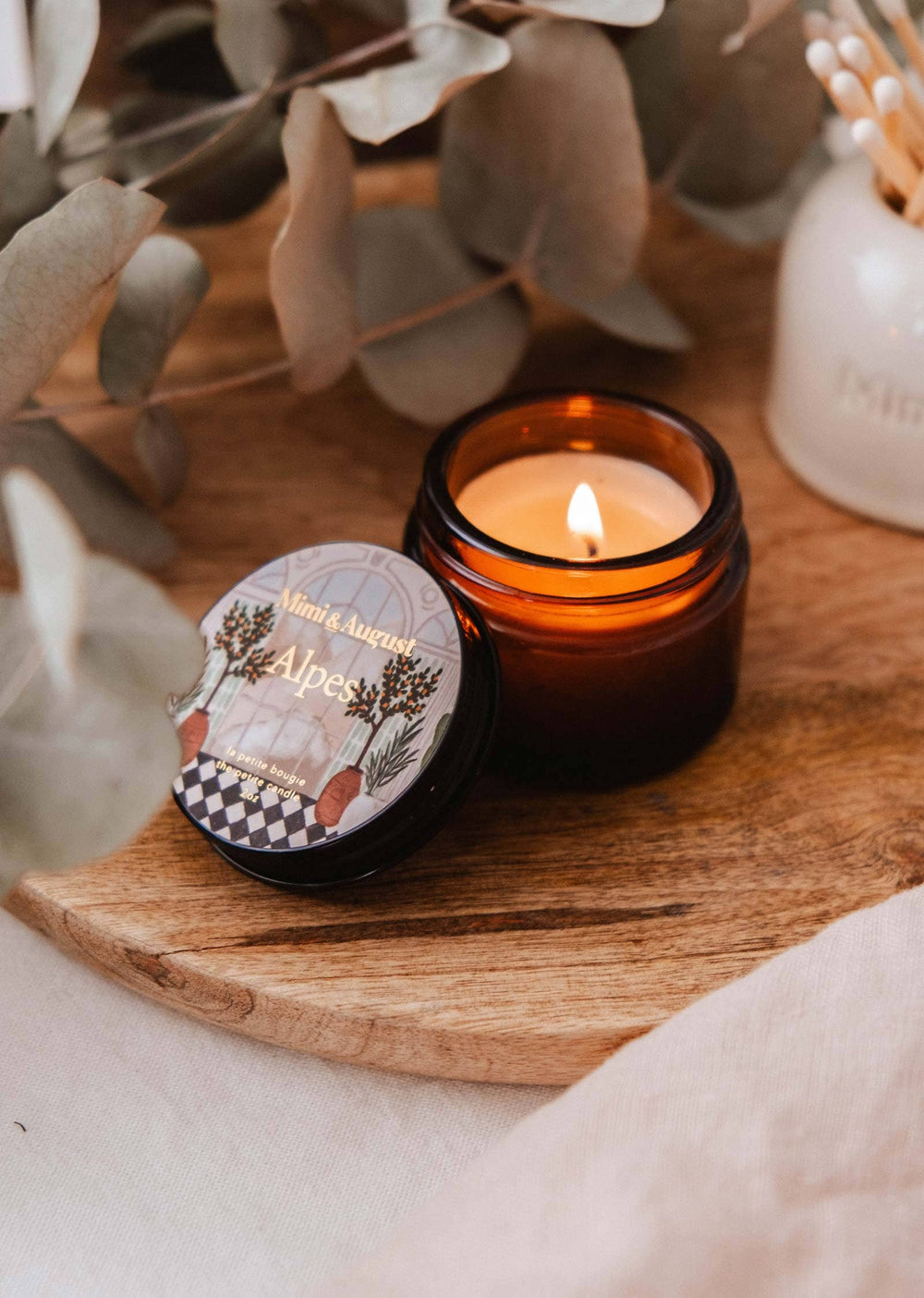 Candle in a glass jar with a decorative lid on a wooden surface, surrounded by eucalyptus leaves.