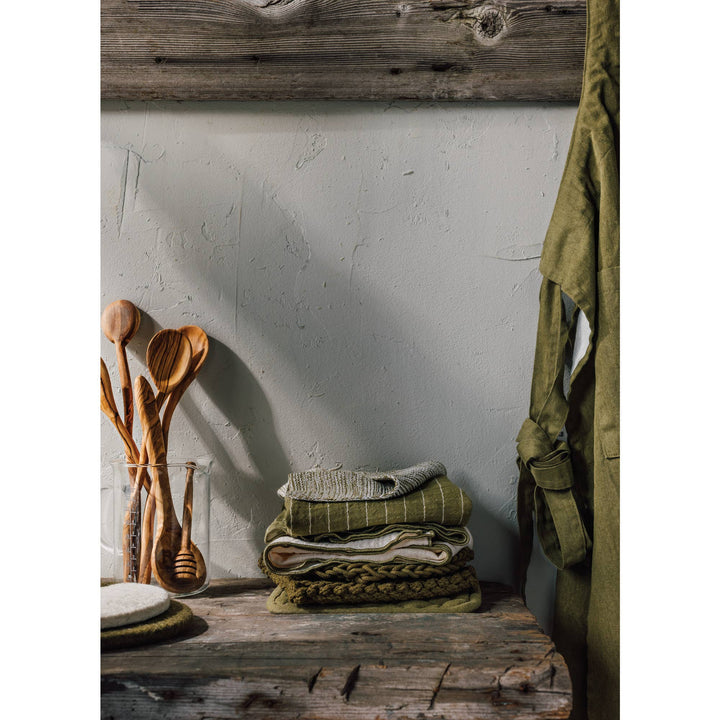 Wooden spoons in a glass jar on a wooden surface with green towels and a green jacket against a white wall.