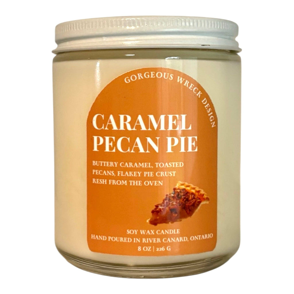 An 8 oz soy wax candle in a clear glass jar with a white screw-on lid. The candle features a prominent orange label with white text reading 'CARAMEL PECAN PIE' and a small image of a pecan pie.