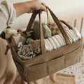 Brown woven basket filled with baby items including stuffed animals, held by a person in a soft, warm-toned room.