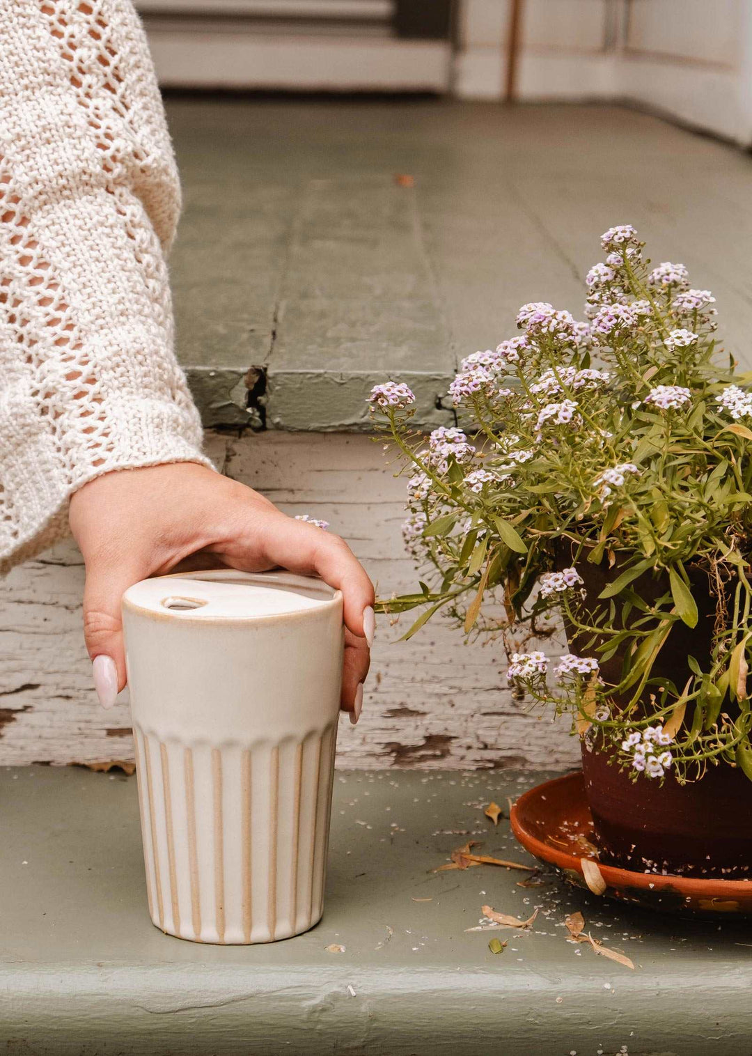 A hand with pink manicured nails holds a cream-colored ribbed ceramic travel mug with a lid. The mug is set on a weathered green wooden surface next to a potted plant with small white flowers.