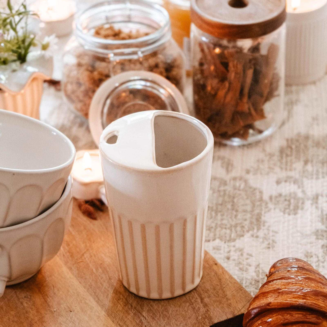 A cream-colored ceramic coffee pour-over dripper with vertical ridges sits on a wooden cutting board, surrounded by coffee beans in glass jars, stacked ceramic bowls, and croissants on a table with candles.