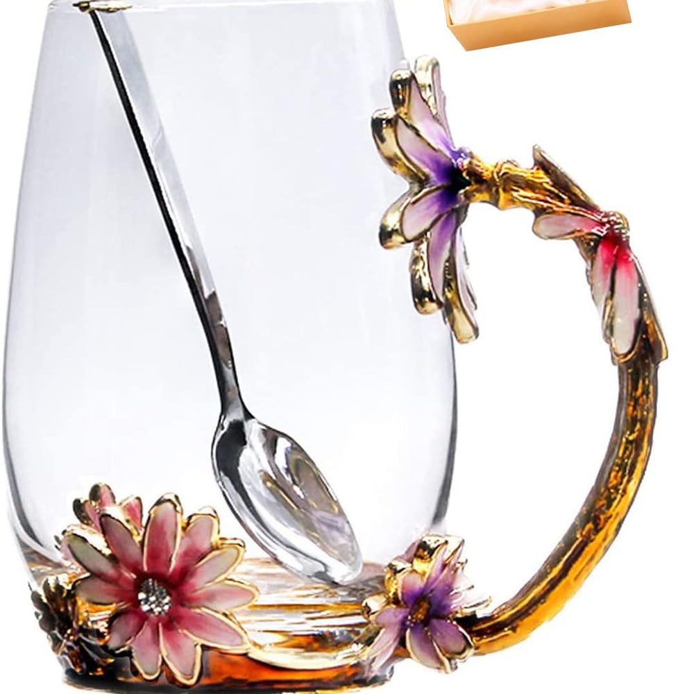 A clear glass mug with an ornate floral handle and spoon, decorated with pink and purple flowers and gold-toned metal accents, shown on a white background with a small gift box in the upper right.
