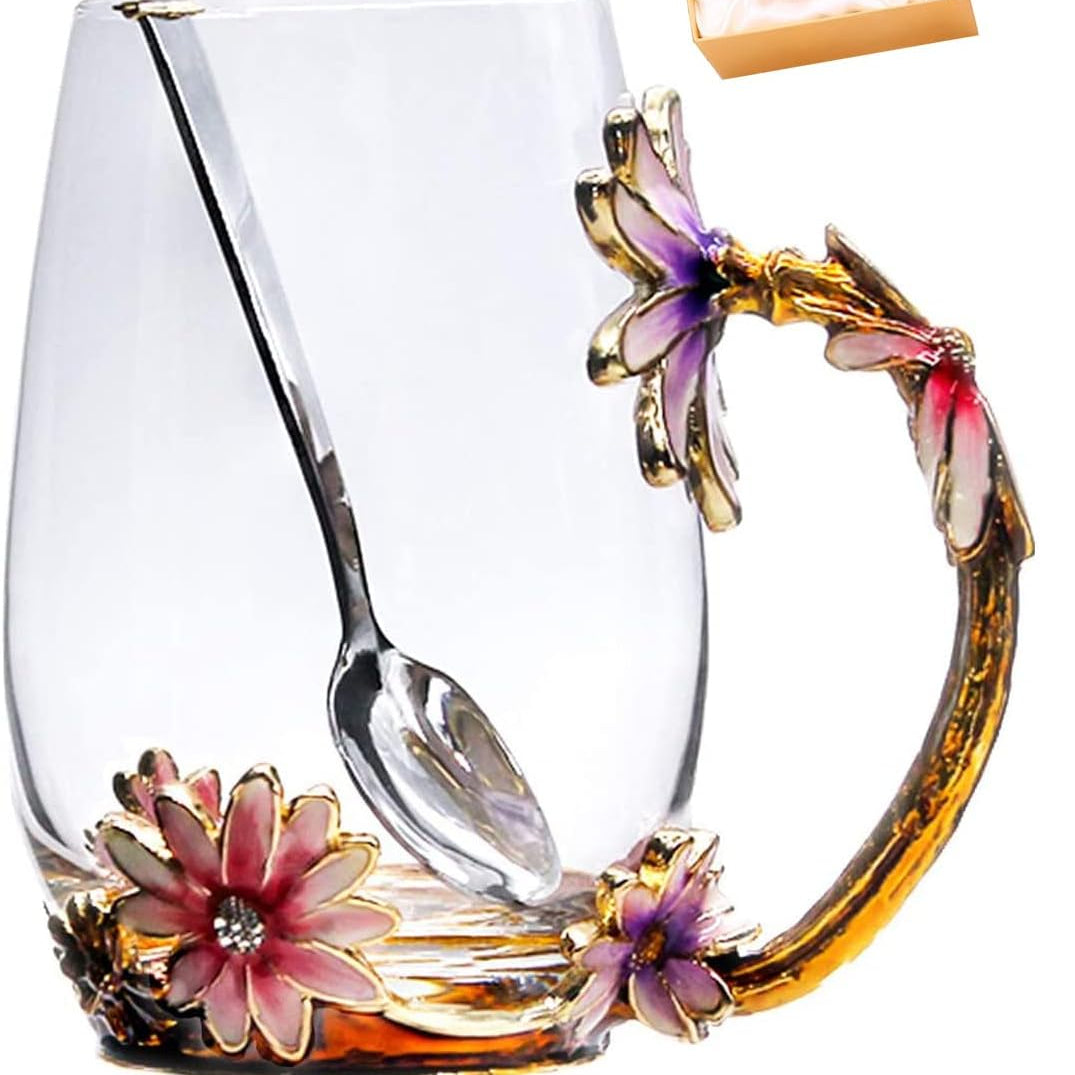 A clear glass mug with an ornate floral handle and spoon, decorated with pink and purple flowers and gold-toned metal accents, shown on a white background with a small gift box in the upper right.