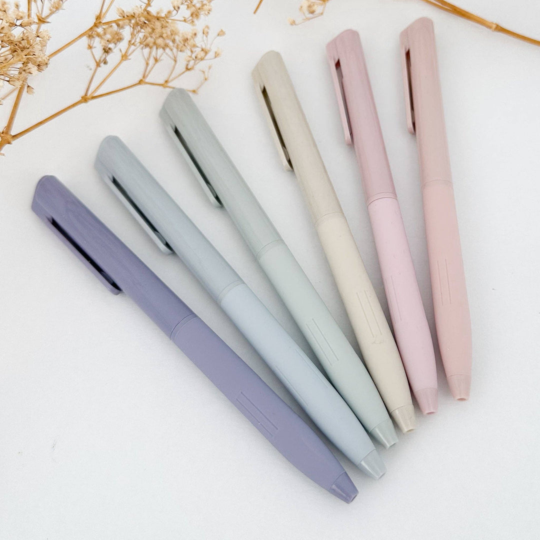 Six pastel-colored ballpoint pens in shades of lavender, light blue, cream, and pink, arranged diagonally on a white surface with dried baby&