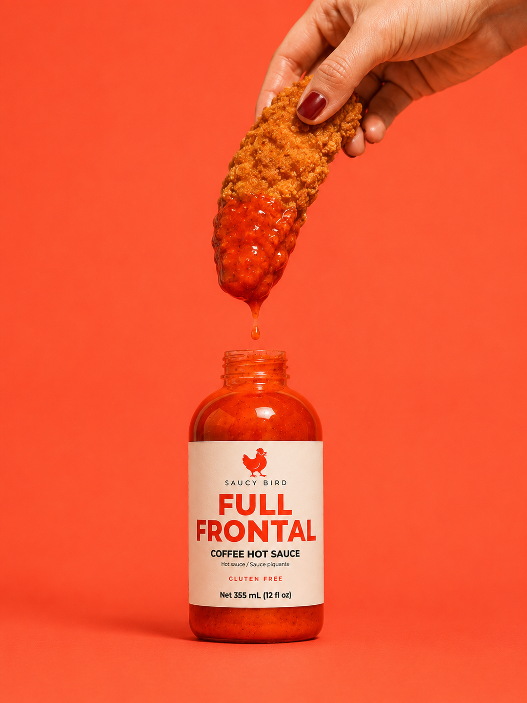 A hand with dark red nail polish holds a breadstick above an open bottle of Saucy Bird Full Frontal Coffee Hot Sauce, with sauce dripping from the breadstick against a solid orange-red background.