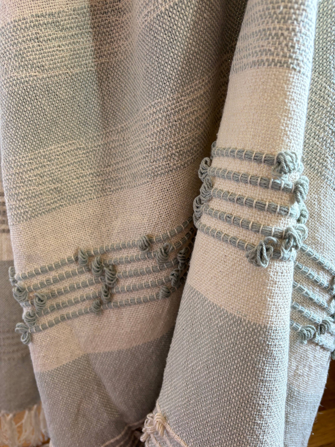 Close-up of a woven throw blanket in soft beige and light gray tones with decorative knotted fringe details along the edge.
