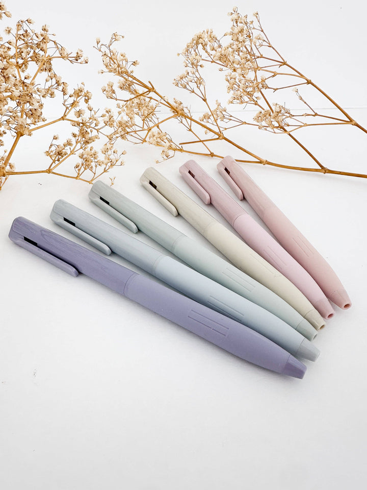 Six pastel-colored ballpoint pens in shades of lavender, light blue, mint, cream, and blush pink, arranged diagonally on a white surface with dried baby&