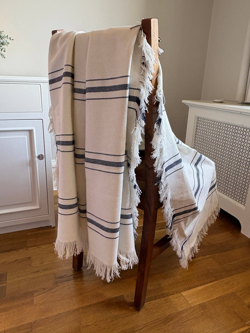 A cream-colored woven throw blanket with navy blue stripes and fringed edges draped over a wooden chair in a home interior setting.