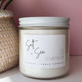 Candle jar labeled 'Salt & Sea' next to a plant on a pink background