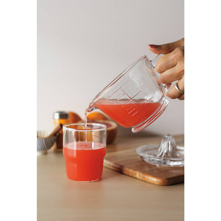 Person pouring red juice from a measuring cup into a glass on a wooden surface.
