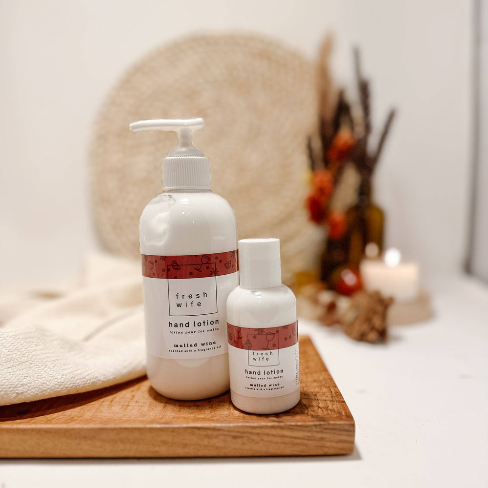 Two white hand lotion bottles from Fresh Wife, a pump bottle and a smaller bottle, displayed on a wooden tray with a folded cream towel and a softly blurred background featuring dried flowers and a candle.