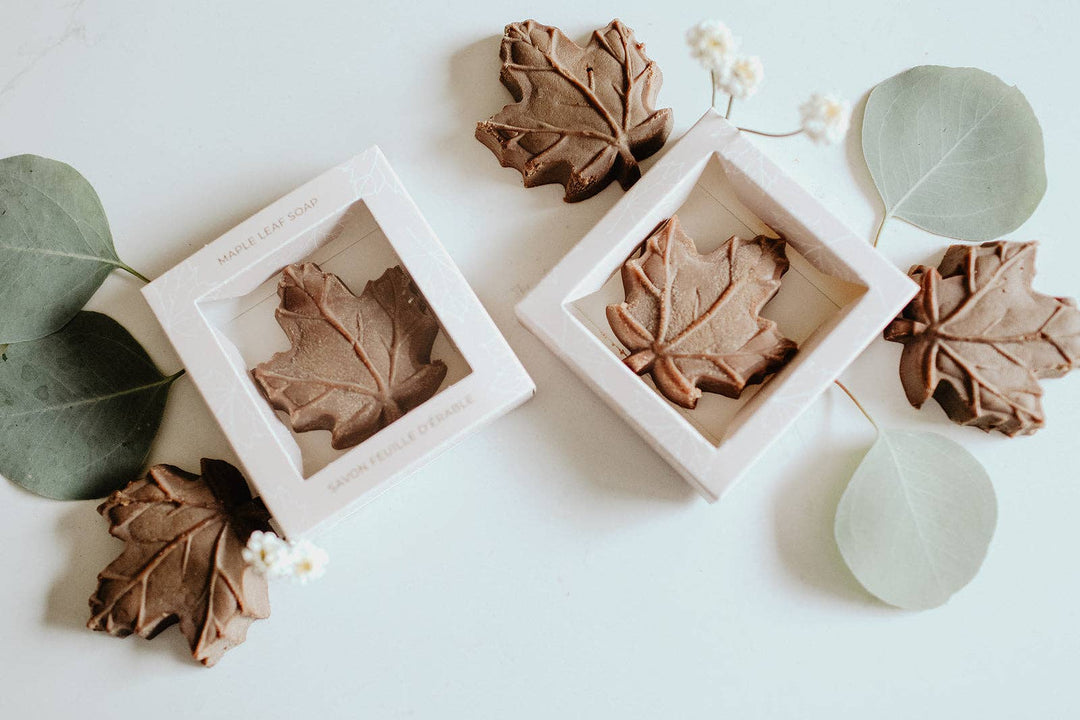 Two square white gift boxes containing leaf-shaped soap bars, arranged on a white surface with decorative eucalyptus leaves and small white flowers. The soap bars are brown and shaped like maple leaves.