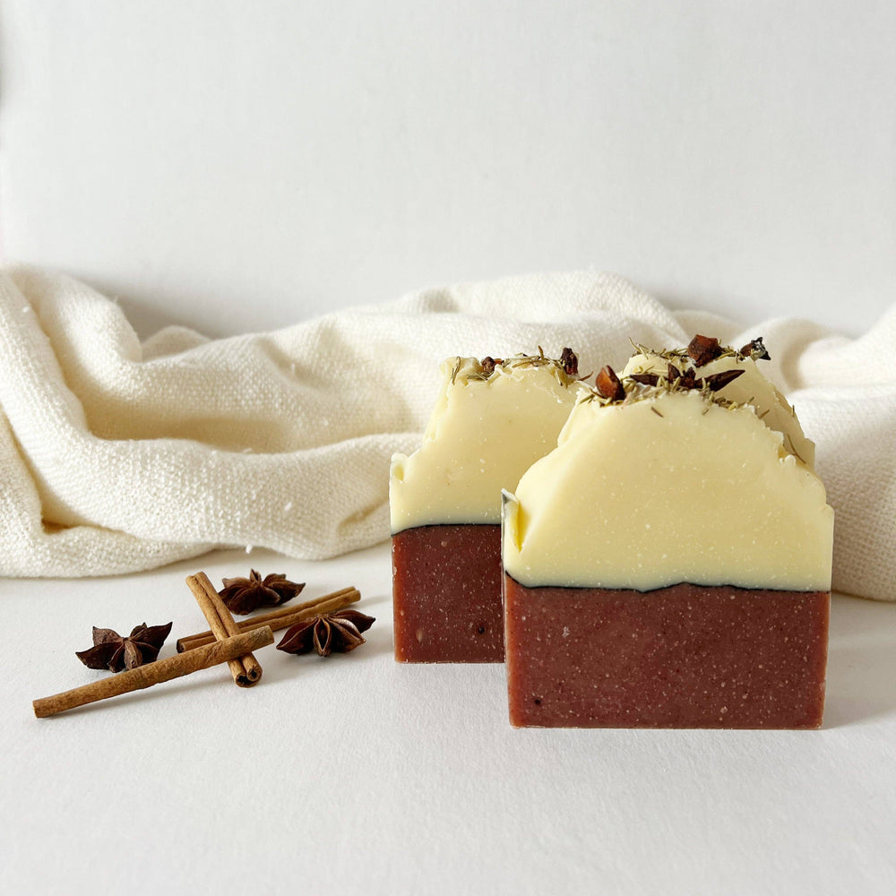 Two rectangular bars of handmade soap with a dark reddish-brown base and a pale creamy top, styled on a white surface with cinnamon sticks and star anise, with a cream-colored textured cloth in the background.