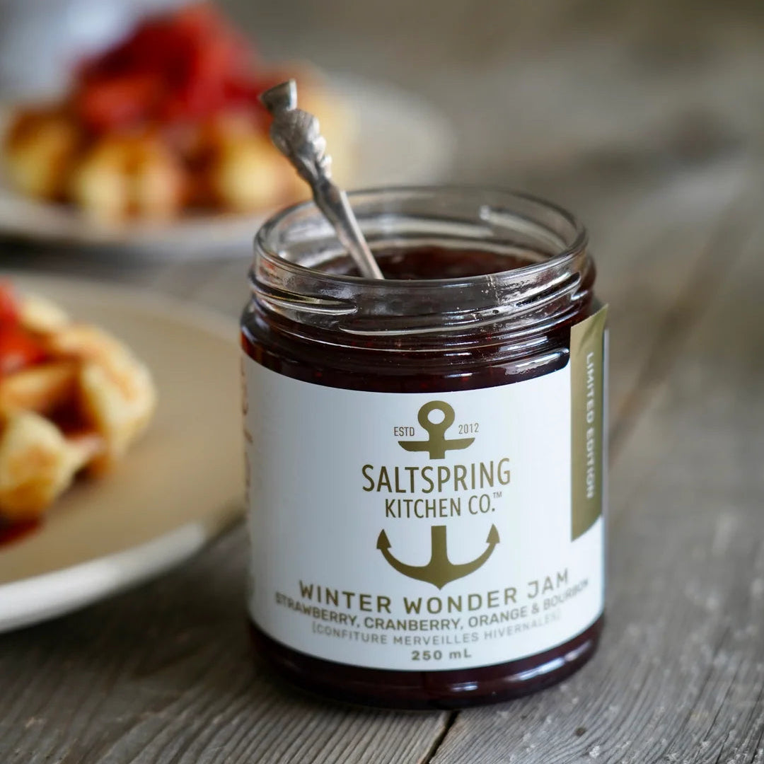 A jar of Saltspring Kitchen Co. Winter Wonder Jam sits on a rustic wooden table with plates of food blurred in the background, indicating a lifestyle setting.