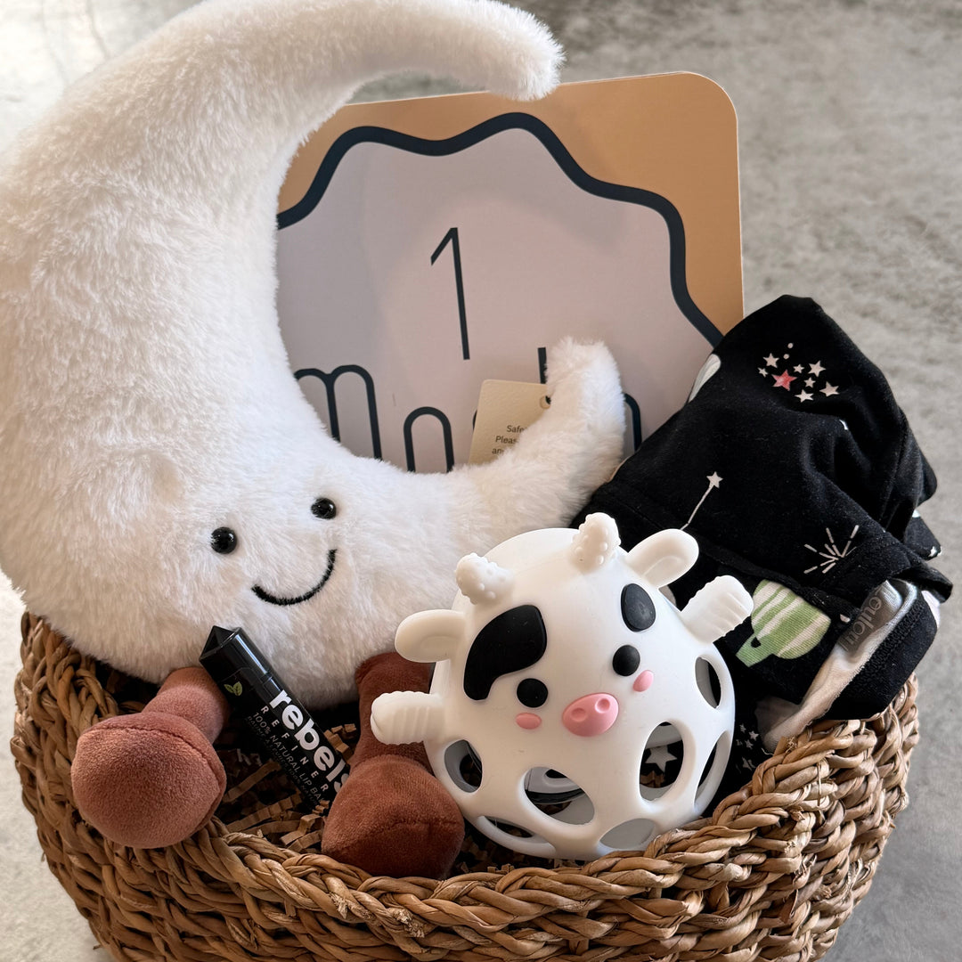 A baby gift basket set in a woven brown basket, featuring a large white crescent moon plush toy, a cow-patterned baby toy, black baby socks, and a wooden 1-month milestone card on a gray concrete floor.