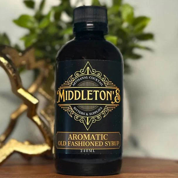 Bottle of Middleton's Aromatic Old Fashioned Syrup on a wooden surface with a decorative heart and plant in the background.