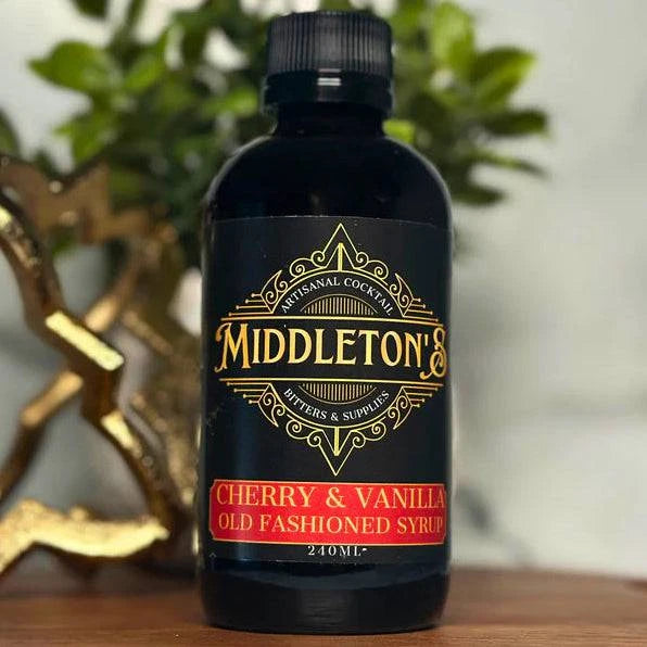 Bottle of Middleton Cherry & Vanilla Old Fashioned Syrup on a wooden surface with a decorative heart and plant in the background.
