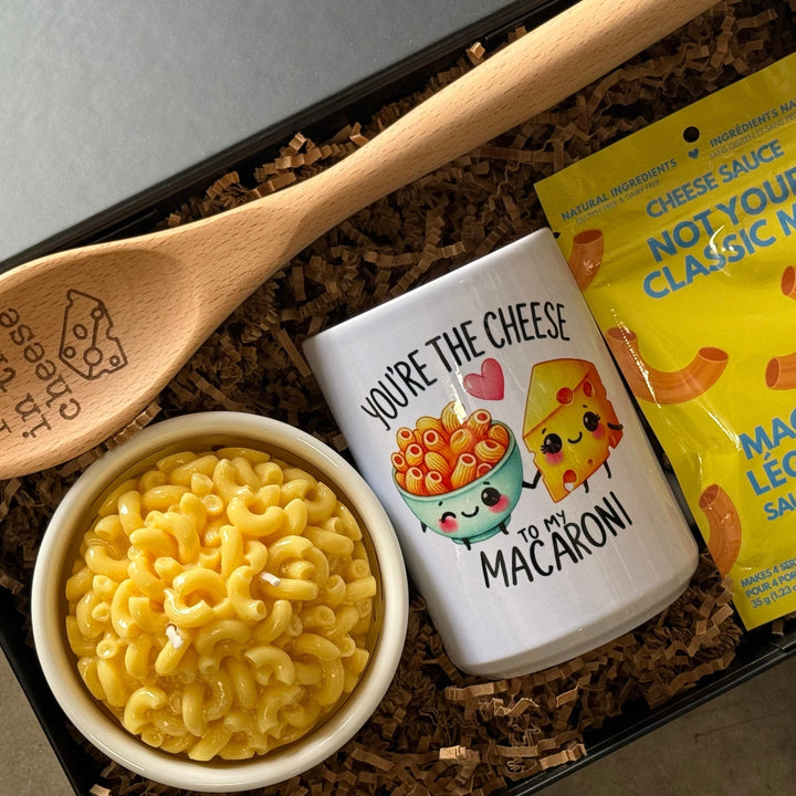 Macaroni and cheese set with a wooden spoon, tumbler, and packaging on a gray surface.