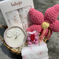 Lulujo baby products including a basket, candle, and flowers on a textured surface