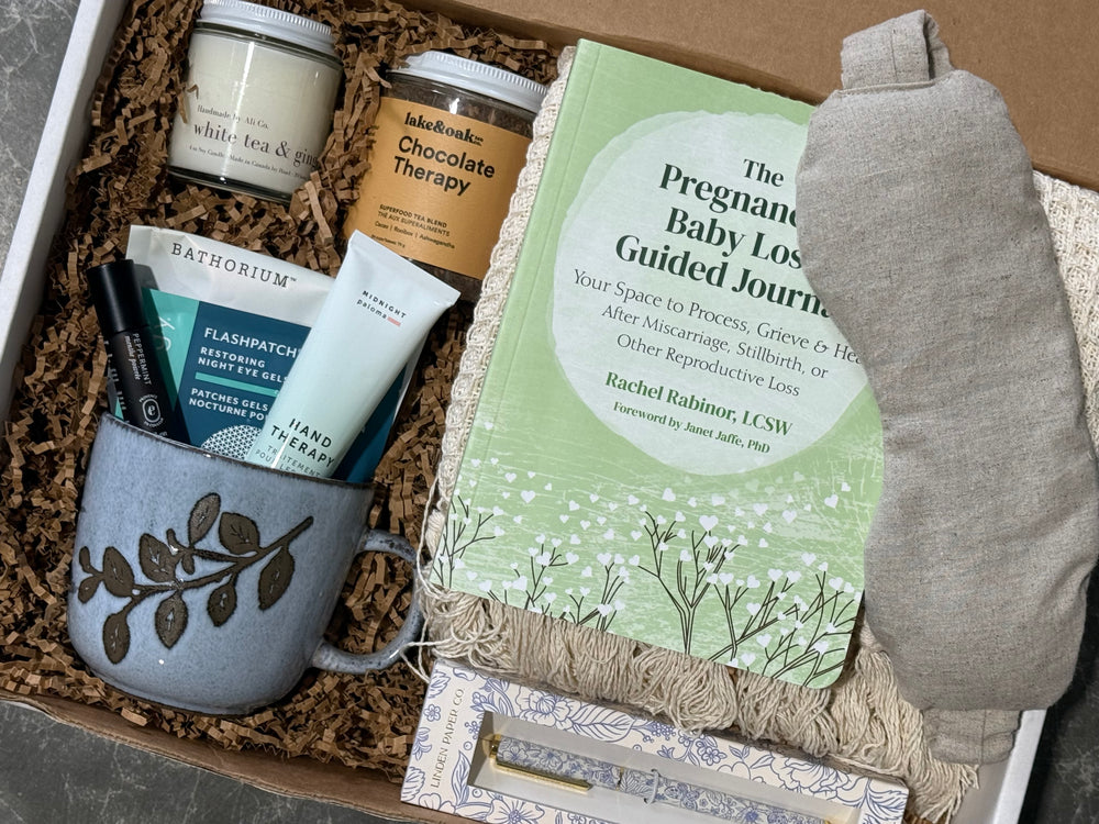 A curated gift box set containing a sage green 'The Pregnant Baby Loss Guided Journal', a gray ceramic mug with a leaf pattern, Bathorium breast cancer support cream, a jar of chocolate therapy, a candle, and a gray fabric pouch, all arranged in brown paper shred.