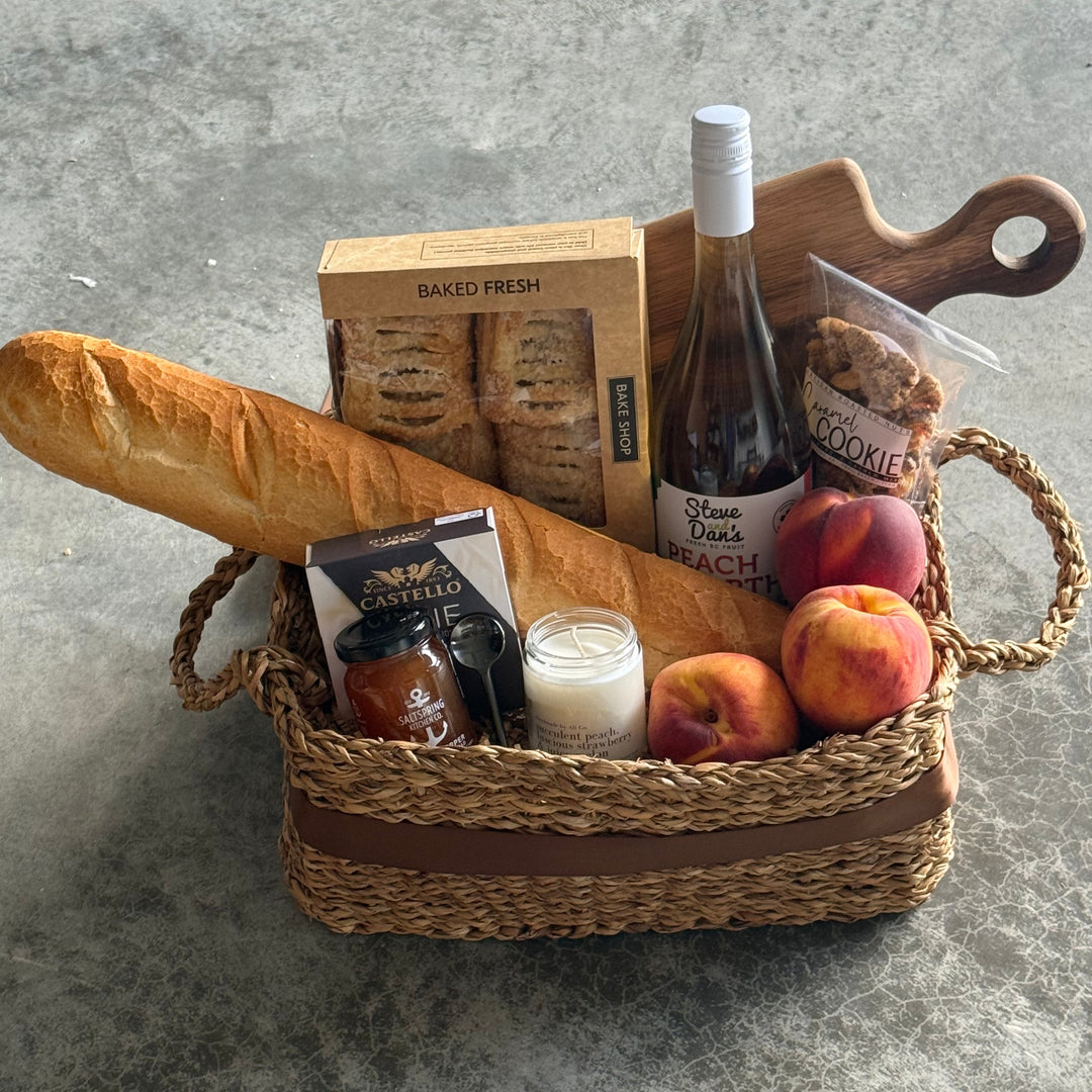 A woven straw picnic basket filled with food items, including a baguette, wine bottle, fruit, and packaged snacks, arranged on a gray concrete surface.