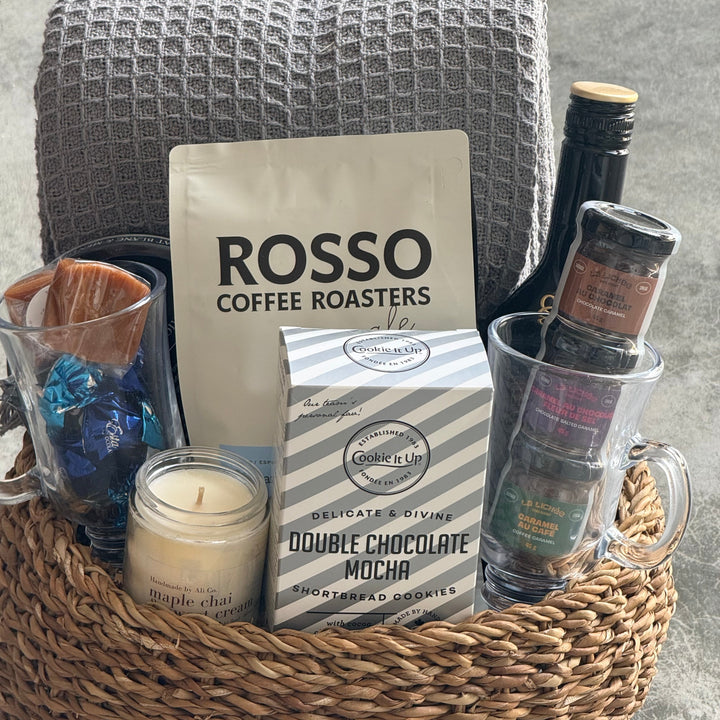 A curated gift basket in a woven natural fiber tray, featuring a bag of Rosso Coffee Roasters, a box of double chocolate mocha cookies, a candle, a gray waffle-knit towel, and assorted packaged treats in glass mugs and a bottle, set on a gray concrete surface.