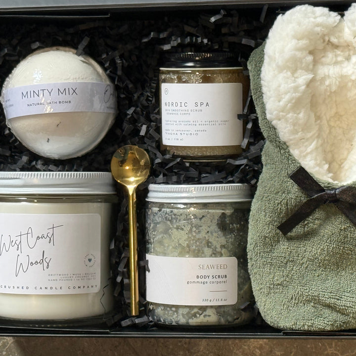 A curated spa gift set in a black box with shredded black paper filler, featuring a round bath bomb labeled &