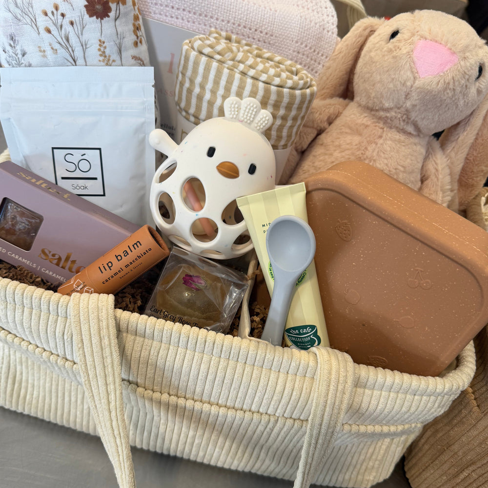 A curated gift basket filled with baby and mom-and-me items, including a plush bunny, a ceramic chick, a lip balm bar, a small soap dish, a storage container, and folded textiles, arranged in a cream corduroy basket on a tabletop.