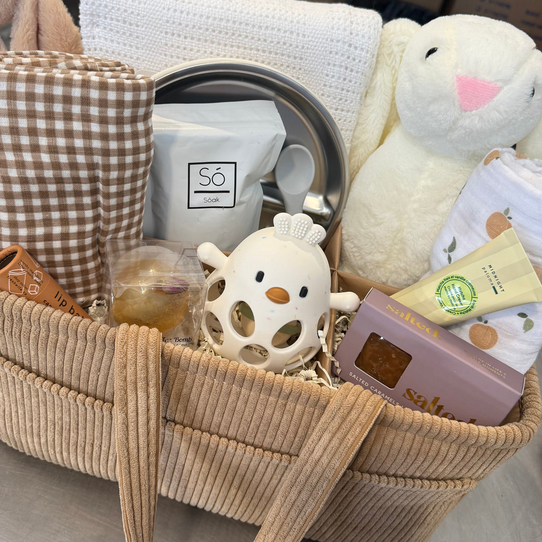 A baby gift basket filled with baby items including a plush bunny, a white knit blanket, a brown-and-white check blanket, a white cloth towel, a small white duck-shaped teething toy, a metal cup, and packaged baby accessories, set on a tabletop with boxes in the background.