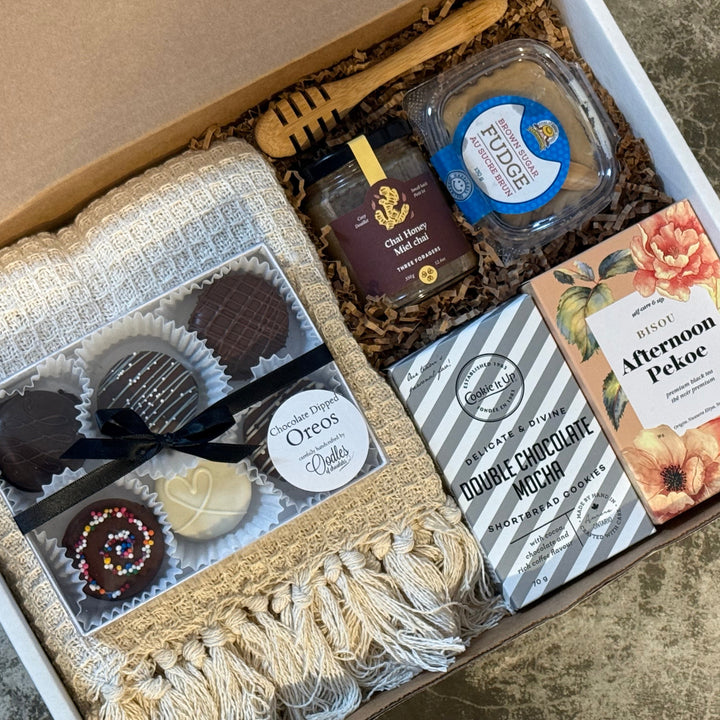 A curated gift box filled with assorted treats and items, photographed from above on a stone-like surface. The box contains chocolate truffles, a honey dipper, a jar of chile honey, a packaged food item, a striped chocolate bar, and a floral card or booklet.