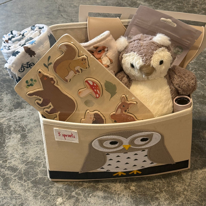 A baby gift basket set on a gray stone surface, containing a beige fabric storage basket with an owl design, a plush owl toy, wooden animal puzzle pieces, and a folded patterned blanket.