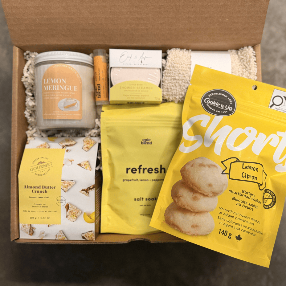 Open box containing various items including a candle, cookies, and snacks on a neutral background