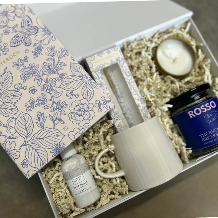 Gift box with a floral notebook, candle, mug, and jar of tea on a wooden surface.