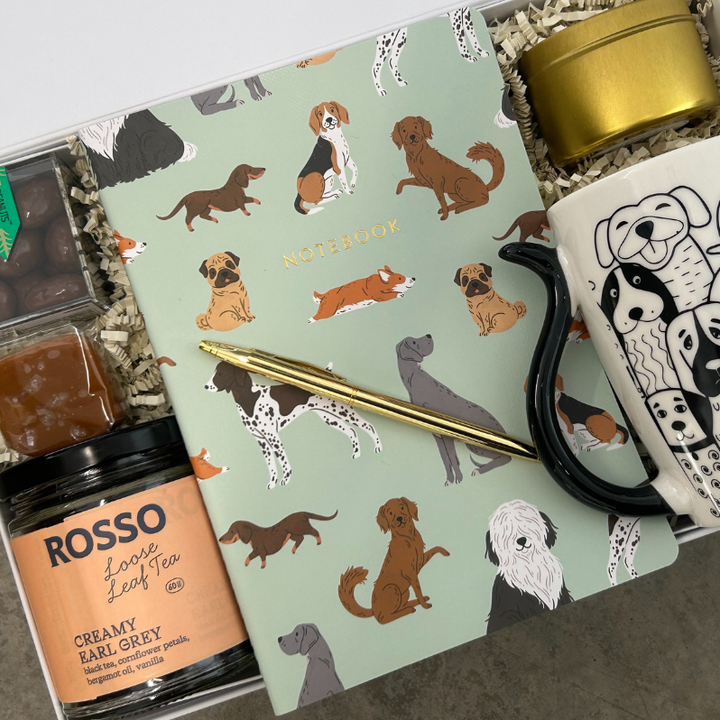 Gift set with tea, chocolate, and a mug with a dog illustrated notebook on a light gray background.