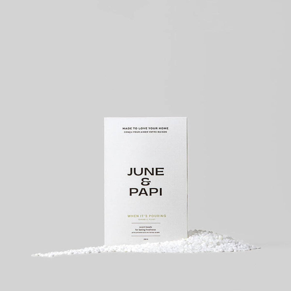 A minimalist white product box with black text reading 'JUNE & PAPI' stands upright on a light gray background, with a small pile of white crystalline material spread along the bottom in front of it.