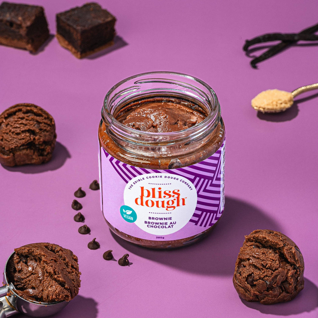 Jar of Bliss Dough Brownie batter with brownies and chocolate chips on a purple background