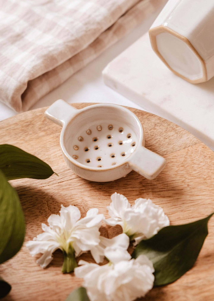 White ceramic strainer on a wooden surface with white flowers and green leaves.