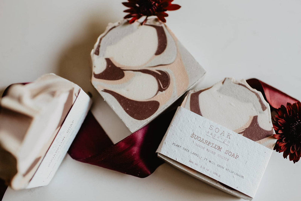 Three artisanal marbled soap bars in decorative paper and fabric wraps, styled with dark burgundy flowers on a clean white background. One soap bar is labeled 'SOAK MALIBAR SUGARBELIN SOAP'.